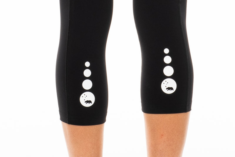 Close view of reflective logo on calf of black leggings. Mid-calf length leggings.