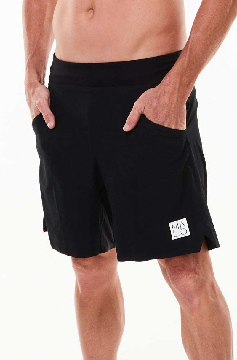 Left view model wearing Arvo Shorts with hands in pockets. Black running shorts with reflective logo on left thigh.