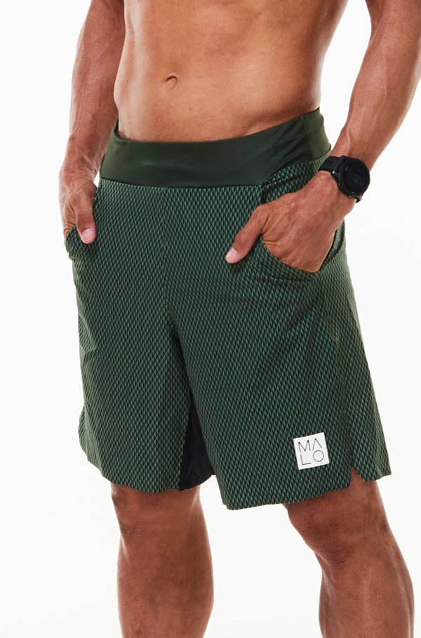 Left view model wearing Arvo Shorts with hands in pockets. Green running shorts with reflective logo on left thigh.