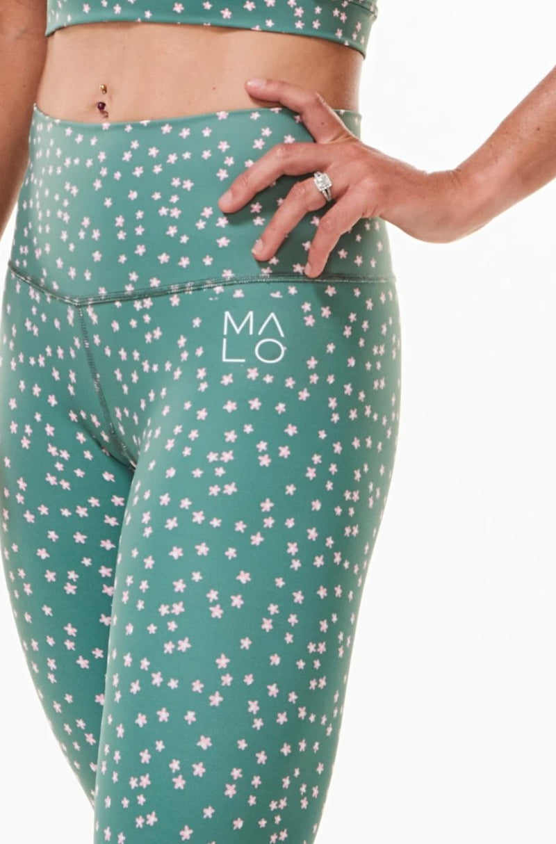 Close view left hip of Hi Rise Luxe Leggings. Green leggings with daisy print and white 'MALO' logo.