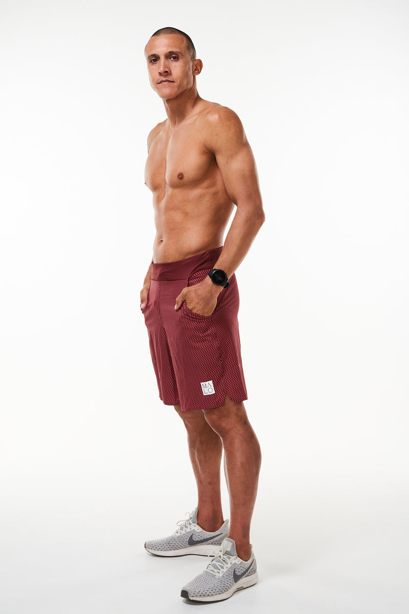 Left view model wearing Arvo Shorts with hands in pockets. Red running shorts with reflective logo on left thigh.